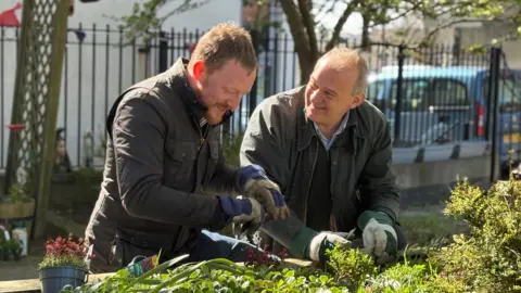 Ed Davey has short grey hair and is wearing a khaki jacket over a black jumper. He is standing next to Newcastle Lib Dem leader Colin Ferguson as they plant flowers in a memorial garden in Heaton, Newcastle. Colin Ferguson is wearing a khaki jacket and has short brown hair. Both men are smiling as they talk to each other. 