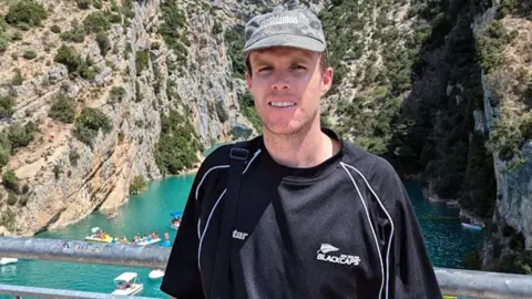 A man wearing a cap and a black New Zealand jersey poses for a photo in front of a canyon filled with turquoise water 