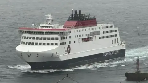 BBC The Manxman ferry, a tall white ship, with red detail on the top and dark base, driving through waves at sea. A buoy can be seen on the right and some festoon street lighting in the close foreground on a grey day.