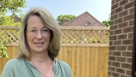 Christine Cullen, wearing a green blouse and a chain around her neck, stands in a garden. She is wearing glasses and has shoulder-length blonde hair. Behind her is a wooden fence and a tree.