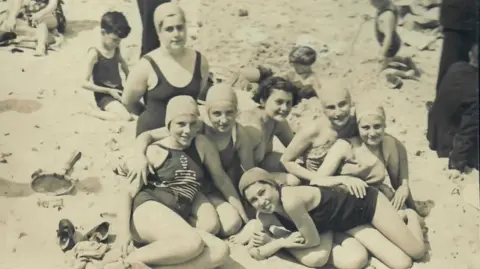 Jason Camis A black and white photograph of seven girls on a beach wearing swimming costumes and caps. They're of varying ages and sitting closely together, smiling. A wooden tennis racquet and some shoes are beside them on the sand. Some boys are digging in the sand behind them. Ilse is lying across the laps of several of the girls and smiling widely.