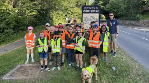 Cucumber PR A group of children who are scouts standing on a grass verge next to a road alongside a scout leader. One of the children is holding a lead with a dog. They are all wearing yellow or orange hi-vis jackets and are standing in front of a sign that reads "welcome to Albrighton"