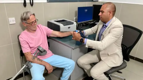 NIKKI FOX/BBC Dr Neil Modha examines a male patient who is in his 60's. Dr Modha has a beige suit on. He is putting a blood pressure cuff on the patient while sitting to the side of his desk with a computer and printer on. The patient has a pink t-shirt and pale blue jogging bottoms. He has glasses on and crutches across his lap.