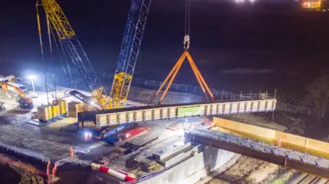 A bird's eye view of the beams of a bridge being lifted into place by a blue and yellow crane at night, under bright lights.