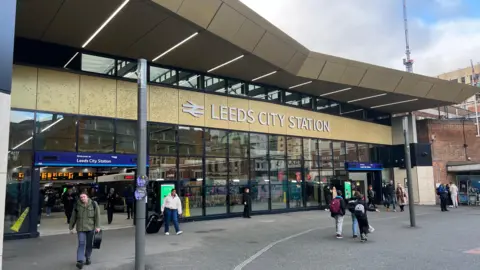 Outside Leeds City station. The building has a glass front, with the name of the station above the glass. It is a sunny day and people are walking in and out of the station. 