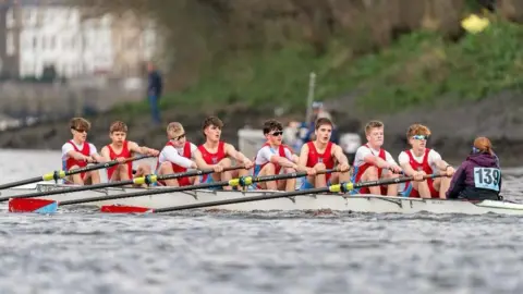 Wallingford Rowing Club 8 young men are pictured in a rowing boat, mid-race. All are wearing red shorts and vests, with some wearing a white top underneath and others wearing sunglasses. A Cox with a purple hoodie is pictured at the rear of the boat. They are all reaching forward with their oars in preparation for a stroke.