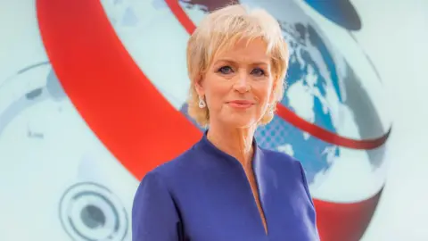 Sally Magnusson in a blue dress standing in front of the BBC News logo in a studio. The red, white and blue news globe can be seen behind her. She is smiling.