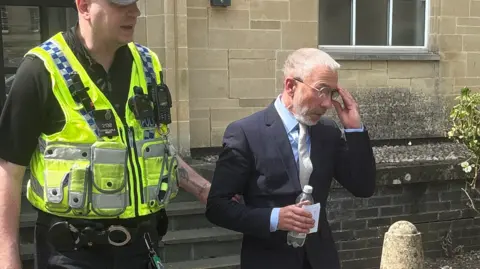 PA Media James Holder is excorted from Cirencester Magistrates' Court by a police officer. He is wearing a dark suit and grey tie
