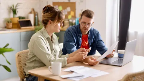 Getty Images Couple go through finances on the kitchen table with bills, a laptop and a mug in front of them.