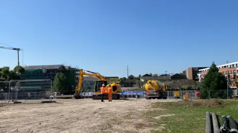A construction site with yellow digger and a truck and two people wearing orange high-visibility suits. 