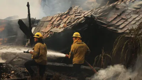 Fire fighters in Malibu hose down a fire. In the background, there is the remains of a burned-down home 