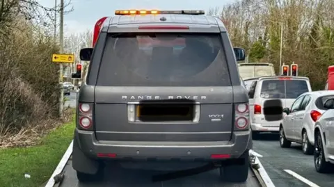 A grey Range Rover, with its licence plate blacked out, on the back of a tow truck at the side of grass verge by a busy road