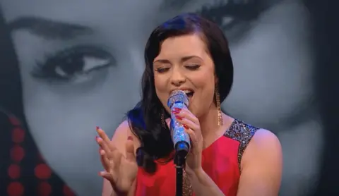 Shona singing close to a glittery microphone with her eyes closed, wearing a sleeveless red dress.