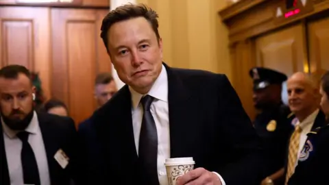 Tesla CEO Elon Musk, Co-Chair of the newly announced Department of Government Efficiency (DOGE), arrives on Capitol Hill on December 05, 2024 in Washington, DC. He is wearing a dark suit and tie and white shirt, is carrying a coffee cup and has a slight smile on his face. 