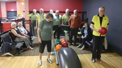 Peter Jordan A group of retired people playing ten pin bowls, one man is wearing a yellow t-shirt and holding a bowling ball. 