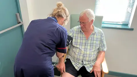 Un hombre de pelo gris, que lleva gafas y una camisa a cuadros azules y verdes, está siendo sometido a una medición de presión arterial por una enfermera.