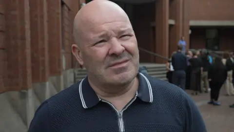 Nigel Hotham standing outside Hull Crown Court. He has a shaven head and is wearing a blue and white polo shirt.