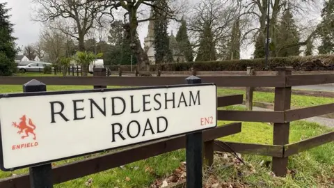 Road sign of Rendlesham Road, with the Enfield emblem on it, in front of a brown fence