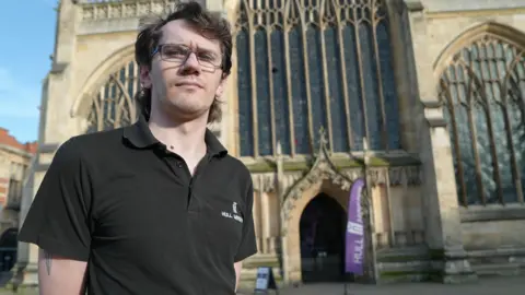 Jasmine Lowe/BBC News James Cameron - a man in a polo neck top with glasses and light brown hair. He looks frustrated and disheartened. He is standing in front of Hull Minster with a circular hole high up in the top left area of the stained-glass. 