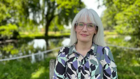 Councillor Heather Delaney sits in front of a large pond at, a the grounds of Nailcote Hall Hotel. Behind her are trees framing the pond. She is wearing a multi coloured shirt, and lilac framed glasses. She has long white hair with a fringe.