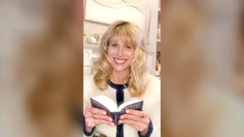 Lucy Punch from BBC series Amandaland holds a dictionary open and smiles. She's wearing a cream cardigan with navy edges.