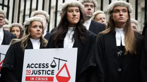 PA Media Three female barristers in white wigs and black gowns, holding a sign which reads Access to Justice in Crisis.