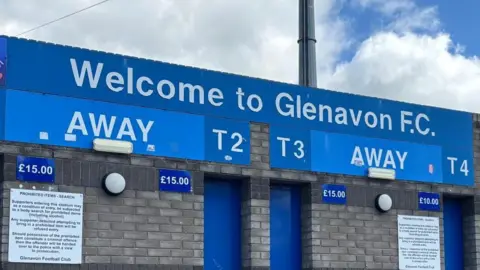 A picture taken outside Glenavon Football Club in Lurgan. There is a 'Welcome to Glenavon FC' sign, with four ticket booths within the grey brick wall.