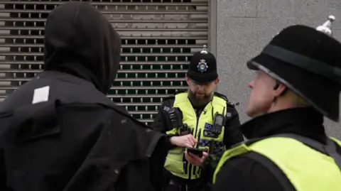 PC Gareth Bielby is in the middle and standing in front of a man dressed in black who has his hood up and Sgt Lee Waller. Gareth is checking his phone, Lee is talking to the man dressed in black.
