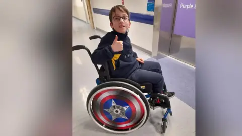 supplied A young boy wearing glasses  spins in his wheelchair and holds a thumb up to the camera.