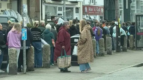 Lucy Sheers A view of a long queue of people on a pavement next to a row of shops. 