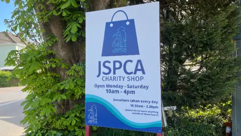 Outdoor sign for a JSPCA charity shop mounted on two red posts among trees. The blue-and-white sign shows a shopping bag logo and reads: "JSPCA Charity Shop. Open Monday–Saturday 10am–4pm. Donations taken every day 10.30am–3.30pm (please do not leave anything outside of these times)."
