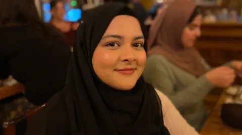 Thahera is wearing a black headscarf and a white jumper, the background is out of focus but another woman can be seen sitting behind her