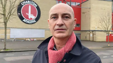 Bald man looks into camera. He is wearing a red scarf. Behind him is the Charlton Athletic club crest