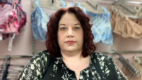 A close up shot of a woman looking directly at the camera. She has short wavy purple hair, just past her shoulders, and has makeup on her face. She is wearing a black and white patterned blouse. Behind her are four rows of bras lined up on a rail.