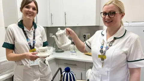 Two women stand in a small kitchen. The women are wearing white hospital uniforms and hospital ID cards around their necks. The woman on the left has brown hair and is holding a teacup. The woman on the right has blonde hair, is wearing glasses, and is holding a teapot.