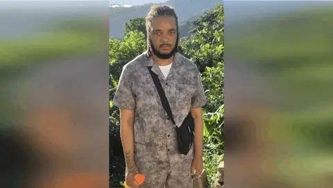 Keanu Taylor wearing a shirt and bag in front of plants. 