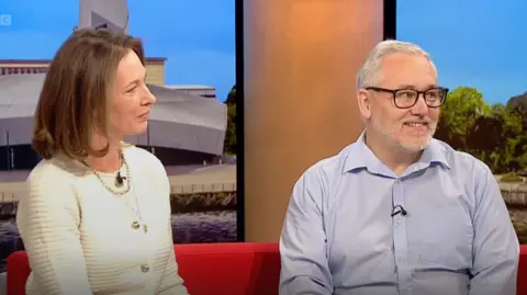 Jinnie Payne and Alan Bowen sitting on the red BBC Breakfast sofa. She is wearing a cream cardigan with gold buttons and a necklace while he wears a blue collared shirt without a tie,