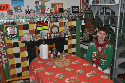 Lisa Watts sits at a small kitchen table covered with a red tablecloth, surrounded by colourful tiled walls, vintage Christmas decorations on the mantel, and shelves filled with festive ornaments.