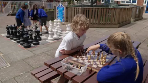 Joe Bilton Two pupils are sat on a brown bench in a school playground playing chess on a wooden board. One girl has light blonde hair and is playing white, she is making her move. Opposite her is a young boy with long blonde hair playing with the brown pieces. Behind them is a set of large chess pieces on the school playground, four children are moving the pieces. 