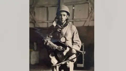 Danny Mason will distribute black and white photographs of Danny Mason right after World War II. He is wearing a parachute regiment uniform. He wears a stiff helmet and a parachute harness over his uniform and holds a gun. He stands in what appears to be a hangar or a shed.