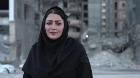Jack Garland / BBC A woman looks directly at the camera. She is wearing black clothes including a headscarf. 