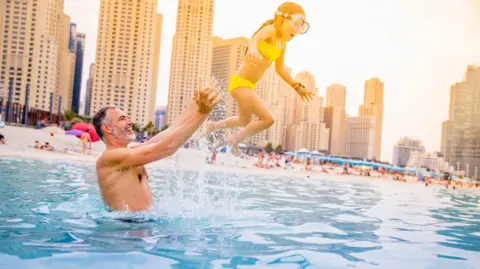 A man in the water at the beach in Dubai throwing his young daughter in the air. There are skyscrapers in the background