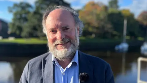 Julian Brazil, Liberal Democrat leader of South Hams District Council, wearing a blue shirt and jacket, standing by the quay in Totnes