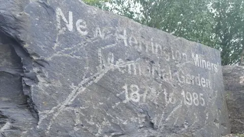 Grey memorial stone in front of trees with faded lettering that says New Herrington Miners’ Memorial Garden 1874 to 1985.