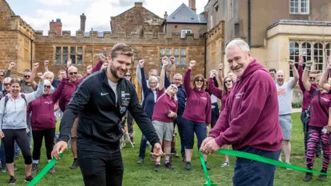 Kirsty Edmonds Two men cut a green ribbon at Delapre Abbey in Northampton. In the background, a large group of people raise their fists in apparent celebration.