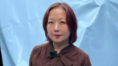 A Japanese woman stands in front of a light blue fabric backdrop with hands behind their back. She is wearing a brown long-sleeved blouse with a bow-tie neckline and dark trousers