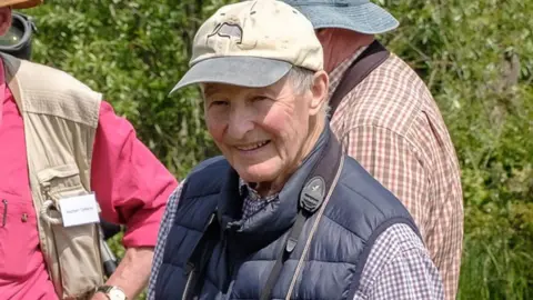 Kingfishers Bridge A head-and-shoulders shot of Andrew Green, standing outside. He is wearing a cream baseball cap with a grey brim and is cleanshaven with short grey hair. He is wearing a blue gilet over a checked shirt. He is smiling. Behind him are two other men and beyond them is shrubbery.
