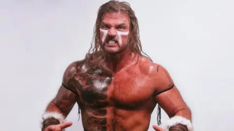 Johnny Jupiter Johnny Jupiter has long blonde hair, white war paint on his face and a large tattoo on the right of his bare torso. He also has fur-like bracers on his wrists and black straps on his upper arms. He is baring his teeth in an expression of anger.