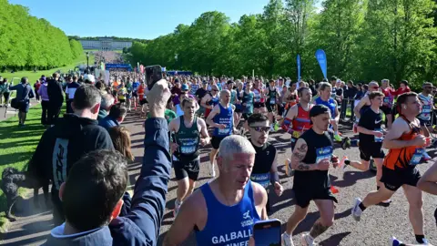 Pacemaker Hundreds of runners exit the Stormont estate which is lined by trees with the parliament building int he distance. A man at the front of the picture holds up his mobile phone to film them