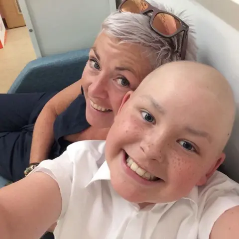 Children with Cancer UK A child with cancer smiles at the camera with his mum behind him